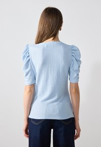 Ljust blått texturerat plagg med puffärm, åtsittande silhuett och rund halsringning. Matchas med mörka jeans.