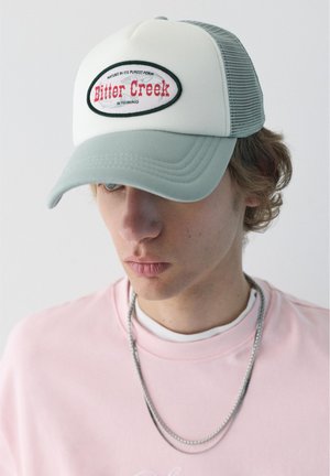 Mladý muž nosiaci svetlosivú a bielu trucker čiapku s logom „Bitter Creek Wyoming“, ružovú mikinu a strieborný náhrdelník.