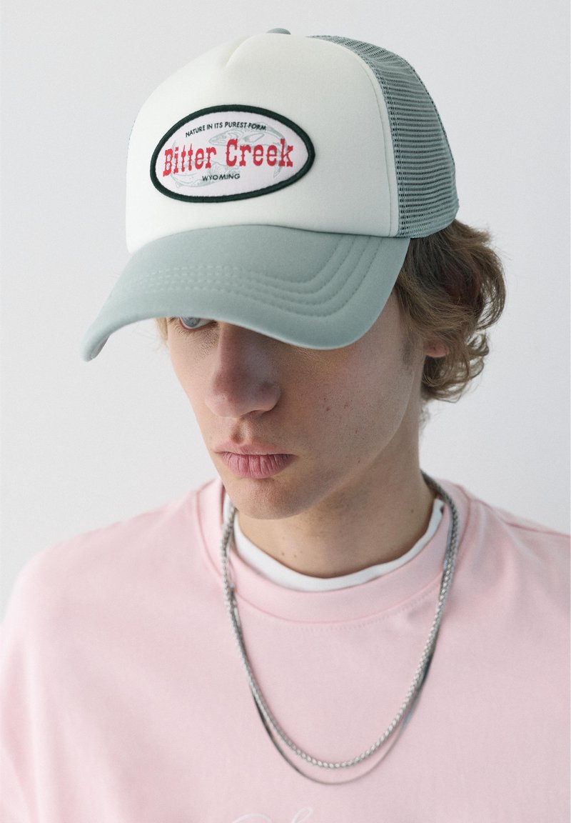 Jeune homme portant une casquette trucker gris clair et blanc avec le logo « Bitter Creek Wyoming », un sweat-shirt rose et une chaîne en argent.