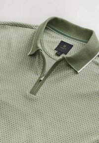 Cămașă polo cu mânecă scurtă, de culoare verde deschis, cu un model tricotat texturat. Prezintă un guler cu fermoar și un eticheta de la "NEXT". Accente minime.