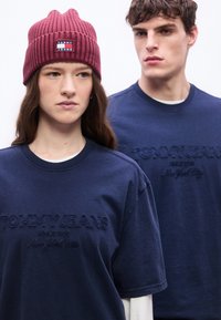 Γυναίκα που φοράει μπορντό σκούφο και μπλε σκούρο πουκάμισο Tommy Jeans στέκεται μπροστά από άνδρα που φοράει το ίδιο μπλε σκούρο πουκάμισο Tommy Jeans.