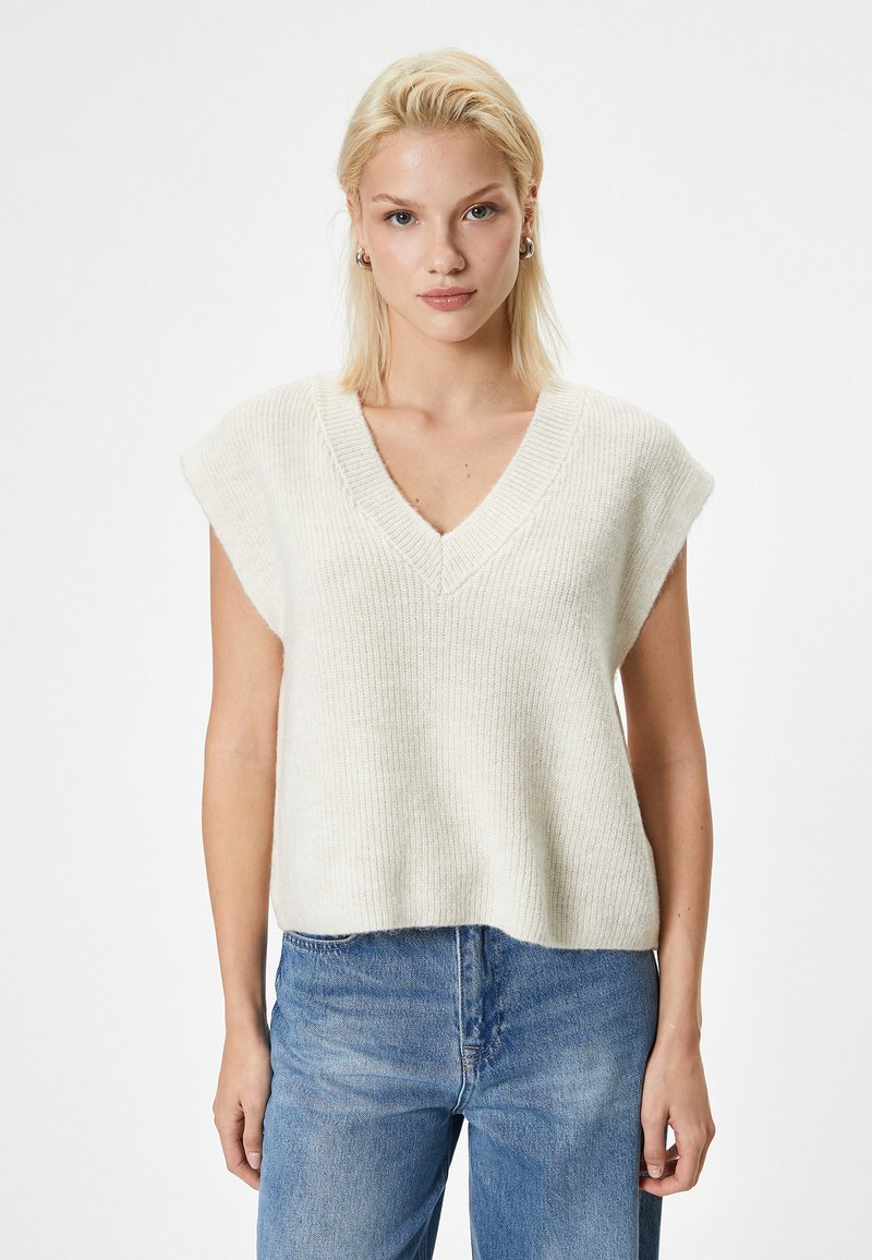 Top a maglia a maniche corte con scollo a V di colore crema, realizzato in un tessuto morbido e testurizzato. Abbinato a jeans blu chiaro, presentato su uno sfondo neutro.