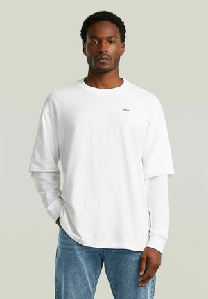 Uomo che indossa maglie bianche a manica lunga e corta sovrapposte con piccolo testo nero sul petto, abbinate a jeans blu, su sfondo semplice.