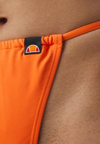 Φωτεινό πορτοκαλί μπικίνι κάτω μέρος κατασκευασμένο από απαλό ύφασμα, με μια μαύρη ετικέτα που διαθέτει ένα πολύχρωμο λογότυπο. Οι πλαϊνές ζώνες είναι λεπτές και ρυθμιζόμενες.