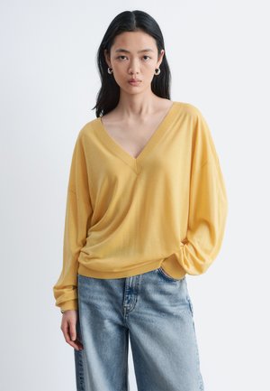 Jonge vrouw met lang zwart haar, gekleed in een gele V-hals trui en lichtblauwe spijkerbroek, staand met één hand in haar zak.