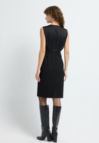 Sort ærmeløs kjole med plisseret ryg, knælængde, med en lille knaplukning bagpå. Sammen med knæhøje, blanke sorte støvler.