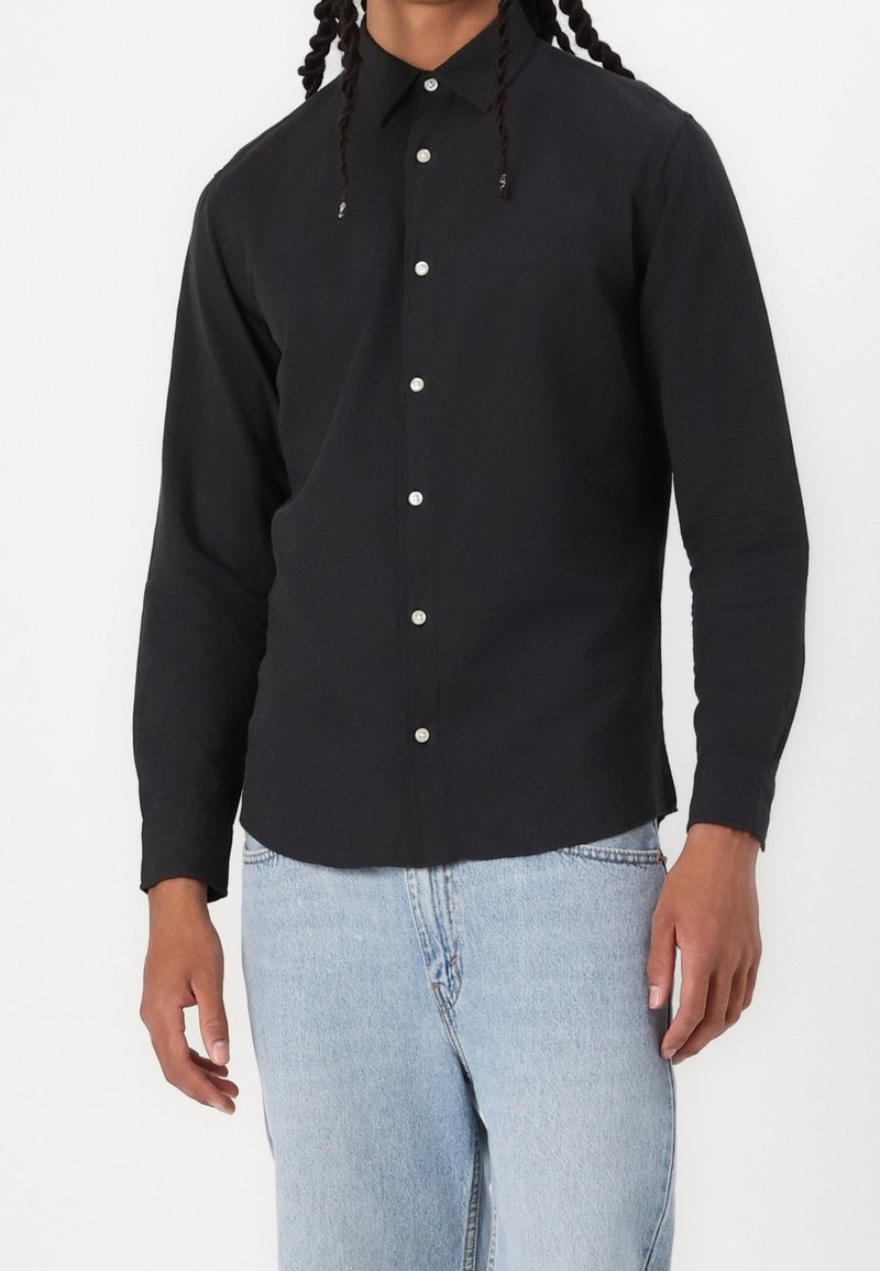 Personne portant une chemise noire à manches longues boutonnée, assortie à un jean en denim bleu clair.