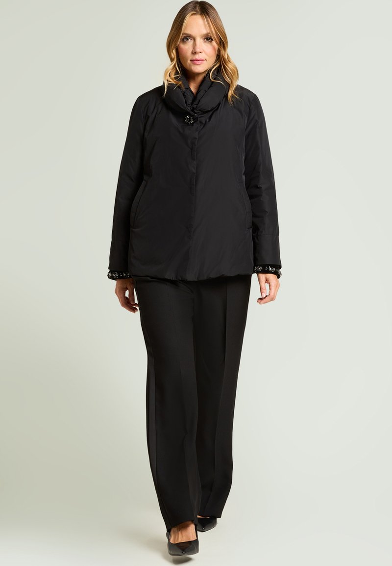 Chaqueta acolchada negra con cuello alto, con cierre decorativo de botones y detalles bordados en los puños, combinada con pantalones negros.