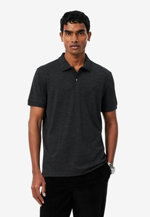 Hombre con una camisa polo de manga corta gris oscuro y pantalones negros, de pie con una mano en el bolsillo frente a un fondo liso.