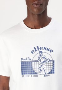 Hvit bomull t-skjorte med et blått grafisk trykk av en tennisspiller og teksten "ellesse Grand Prix TENNIS CLUB" med bakgrunn av rutemønster.
