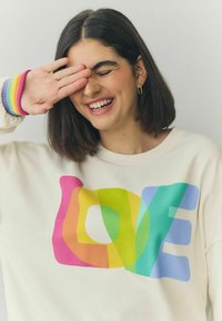 Krémová mikina so zvýrazneným grafickým motívom "LOVE" v pestrofarebných, žiarivých farbách. Rybované manžety v dúhových farbách dodávajú detail.