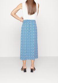 Sukně A-line střední délky z modré látky s bílým květinovým vzorem. Má elastický pas a splývavý střih. Pár černých sandálů na blokovém podpatku.