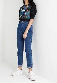 Svart sweatshirt med färgglad blommönster, blå högmidjade jeans och vita sneakers, stående mot en enhetlig bakgrund.