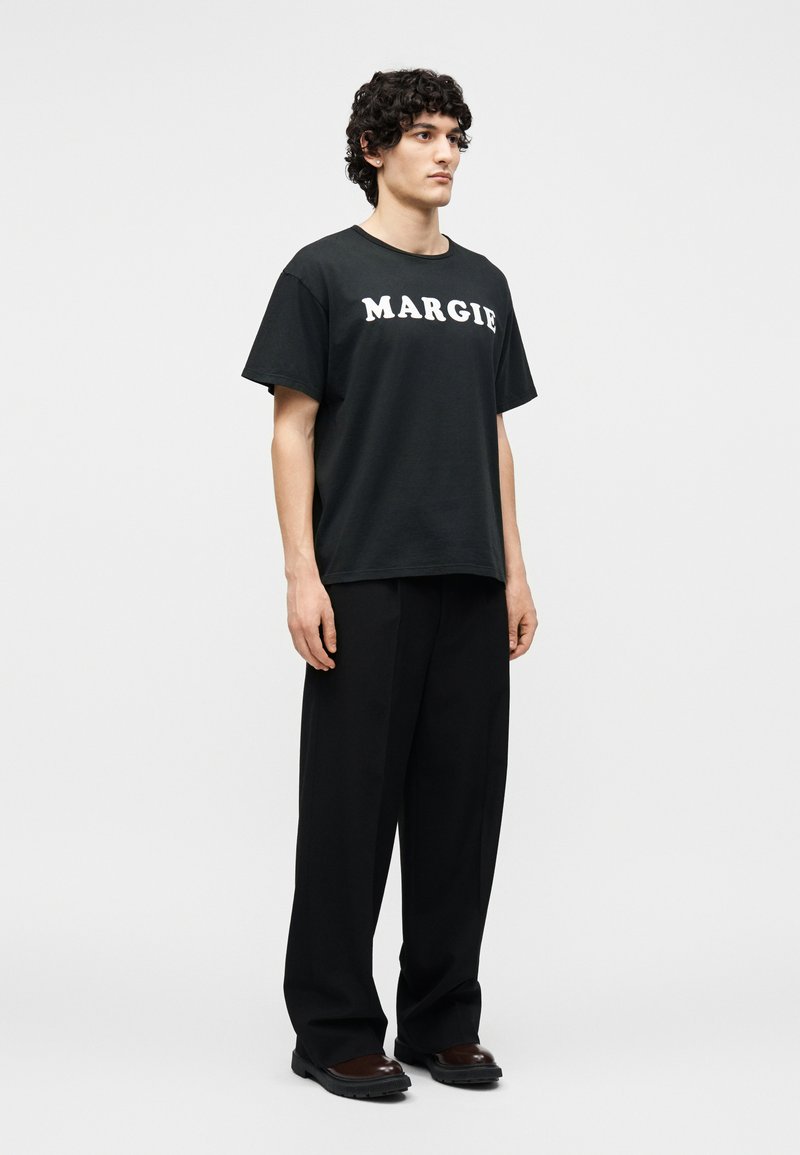Jeune homme aux cheveux bouclés debout, portant un T-shirt noir avec « MARGIE » en lettres blanches, un pantalon large noir et des chaussures marron foncé.