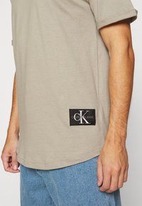 Beige short-sleeve t-shirt with a black Calvin Klein Jeans logo patch near the hem, worn with light blue denim jeans.