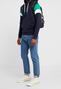 Felpa navy con accenti verdi, bianchi e blu, abbinata a jeans azzurri chiari e sneakers bianche alte. Presenta una tasca frontale e un cordoncino.