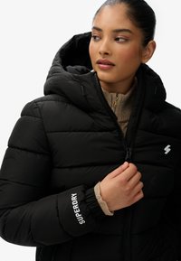 Sort polstret jakke med hætte, med lynlås og elastiske manchetter. Venstre bryst viser et hvidt logo og "SUPERDRY" tekst på ærmet.