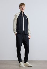 Μαύρο και κρεμ φούτερ με φερμουάρ και μανίκια ραγκλάν, συνδυασμένο με μαύρο παντελόνι φόρμας με τσέπες με φερμουάρ και λευκά αθλητικά παπούτσια.