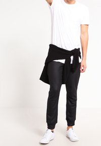 Man som bär en vit t-shirt, svarta joggingbyxor, vita sneakers och en svart tröja knuten runt midjan, stående med en arm lyft.