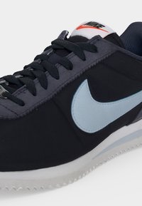 Крупен план на черен и сив маратонка Nike със светлосиня емблема, черни връзки и бяла подметка с текстурирани ръбове.