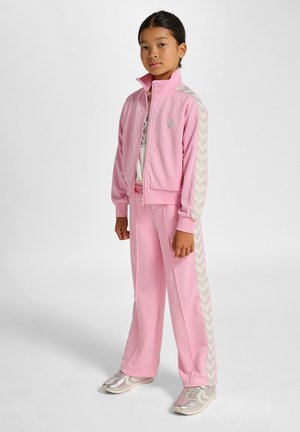 Rosa träningsdräkt med en jacka med dragkedja och matchande byxor. Har chevronmönstrade detaljer på ärmarna och sidorna. Silverfärgade sneakers.