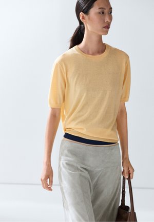 Femme aux cheveux foncés attachés en queue de cheval, portant un haut jaune clair à manches courtes et un pantalon gris clair, tenant un sac à main marron dans une main.