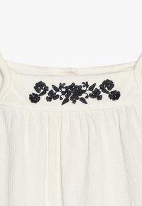 Vêtement blanc sans manches avec broderie florale noire le long de l'encolure et plis verticaux sous la poitrine.