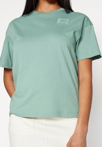 Světlé zelené bavlněné tričko s krátkým rukávem, s malou značkovou nášivkou na horní levé straně a uvolněným střihům.