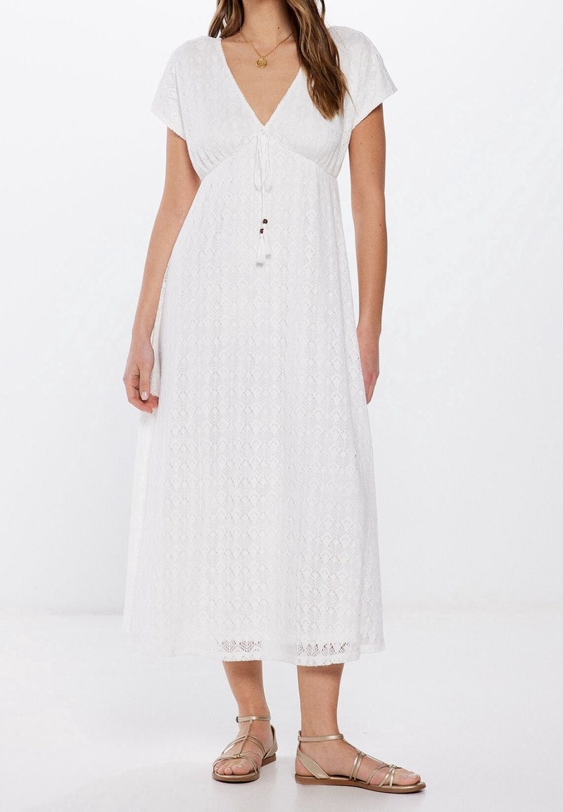 Femme portant une robe midi en dentelle blanche avec manches courtes, col en V et détail noué, associée à des sandales plates dorées à brides.