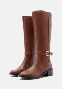 Barna bőr, térd fölé érő csizmák sima felülettel, oldalsó cipzárral és díszítő pánttal, amelyen arany csat található. Tömör sarok kialakítás.