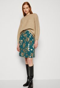 Beige strikkegenser med ribbestruktur, kombinert med en teal blomstrete skjørt med gullmønstre. Modellen har på seg sorte støvler til knærne.