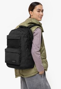 Mochila negra con múltiples bolsillos, tela texturizada y correas ajustables, llevada sobre un chaleco acolchado verde oliva y pantalones de chándal grises.