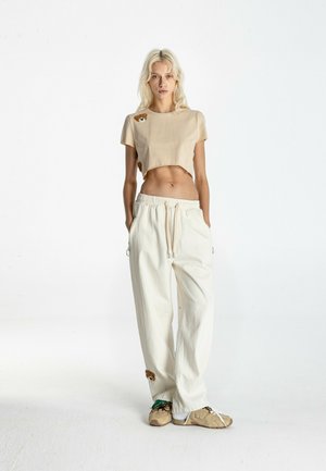 Mujer rubia de pie con las manos en los bolsillos, lleva una camiseta corta beige con parches de osos, pantalones blancos holgados y zapatillas beige.