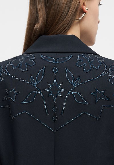 Σκούρο μπουφάν με μπλε κεντημένα λουλουδένια και αστρικά σχέδια στην πλάτη, φορεμένο από ένα άτομο με μακριά καστανά μαλλιά και χρυσούς κρίκους στα αυτιά.