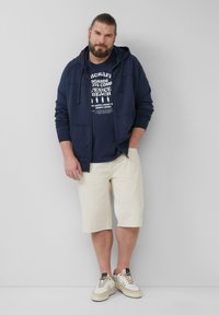 Marine blauwe zip-up hoodie over een donker t-shirt, lichtbeige shorts en beige sneakers met grijze accenten. Soepele stof met een relaxed fit.