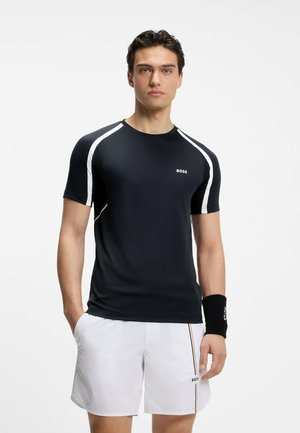 Homme portant un t-shirt de sport noir avec des bandes blanches et un short blanc avec un bracelet noir, debout devant un fond clair uni.