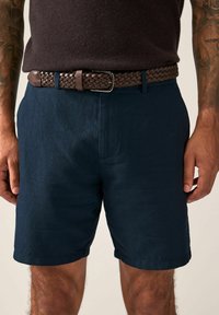 Marineblaue Shorts aus strukturiertem Stoff, mit seitlichen Taschen und geradem Schnitt. Getragen mit einem braunen geflochtenen Ledergürtel.