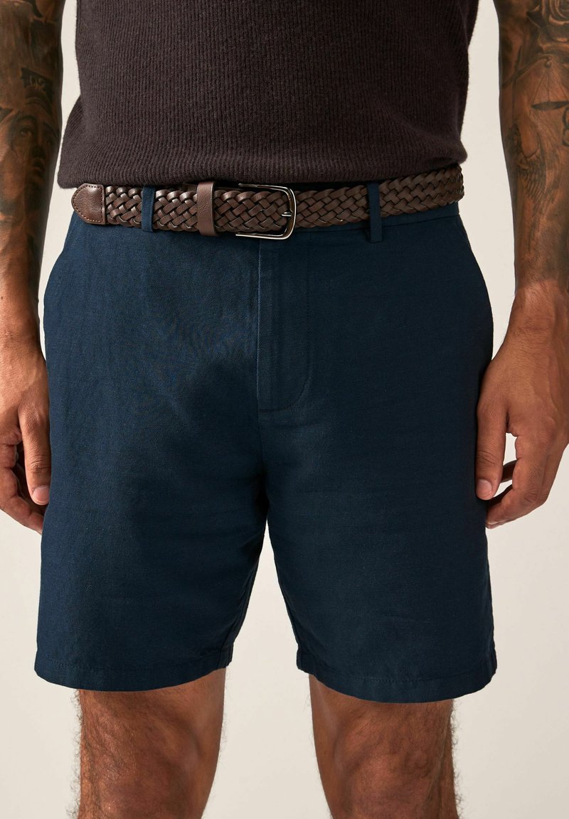 Marineblaue Shorts aus strukturiertem Stoff, mit seitlichen Taschen und geradem Schnitt. Getragen mit einem braunen geflochtenen Ledergürtel.