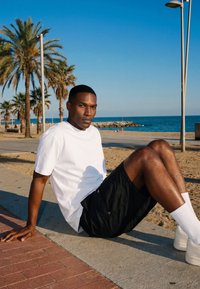 Man in white t-shirt and black shorts sitting on a beach walkway with palm trees and ocean in the background during clear daytime.