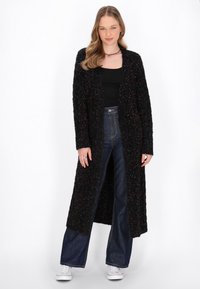 Cardigan lungo nero in tessuto testurizzato, con leggere macchie colorate, apertura frontale e tasche, abbinato a jeans blu a vita alta e sneakers bianche.