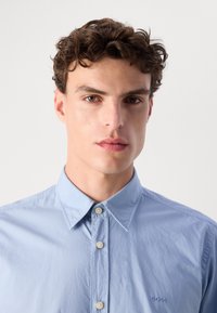 Camisa azul clara de botão com colarinho pontudo, apresentando um tecido texturizado e um logotipo bordado no peito. Os botões são de cor off-white.