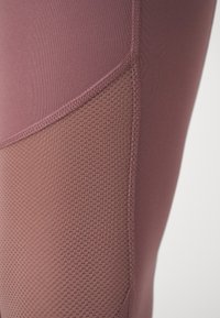 Close-up van mauve leggings met een geperforeerd meshpaneel en versterkte stitching op het been.
