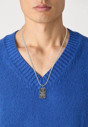 Pull en tricot texturé bleu avec un col en V, porté avec un collier en argent présentant un pendentif rectangulaire avec des détails gravés.