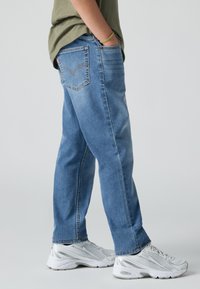 Persona con jeans de mezclilla azules, camiseta verde oliva, zapatillas blancas y plateadas, de pie con las manos en los bolsillos frente a un fondo neutro.