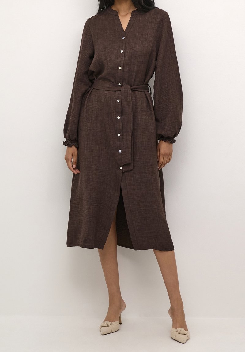Femme portant une robe marron midi à manches longues boutonnée avec une ceinture à la taille et des mules beiges à bout pointu et talons.