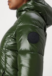 Grön puffjacka med en glänsande finish, som har en logotyp-patch på vänster ärm, huva och sömnad detaljer på tyget.