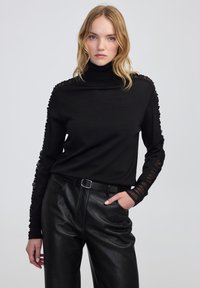 Vrouw in een zwarte coltrui met doorschijnende kantmouwen en zwarte leren broek, staande met één hand in de zak op een grijze achtergrond.