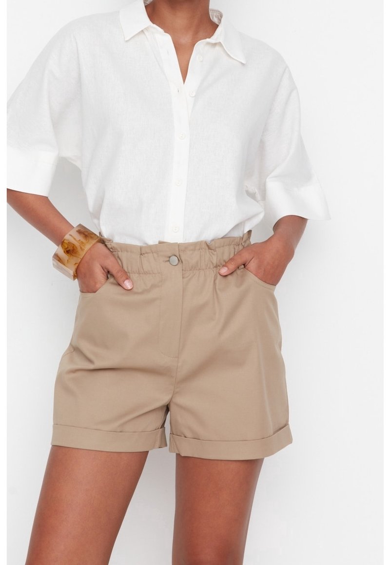 Béžové kraťasy s elastickým pasem a ohrnutými lemy, kombinované s bílou košilí na knoflíky. Outfit obsahuje průhledný náramek.