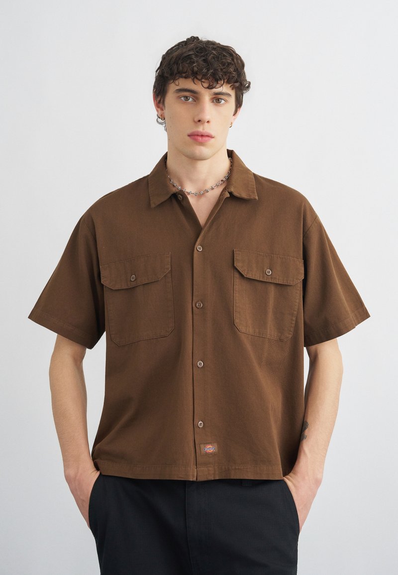 Νέος άνδρας με σγουρά μαλλιά φοράει καφέ πουκάμισο με κοντά μανίκια και κουμπιά, με δύο τσέπες στο στήθος και μαύρο παντελόνι, με τα χέρια στις τσέπες.