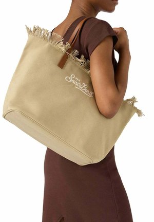 Donna con vestito marrone che porta una grande borsa beige a tracolla con parte superiore frangiata e spalline in pelle marrone.